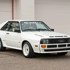 Biały Audi Sport Quattro z białymi felgami i naklejką "Sport Quattro" na przedniej szybie. Ikona rajdów z lat 80.
