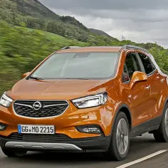 Pomarańczowy Opel Mokka 2015 jedzie po krętej drodze wśród zielonych wzgórz.