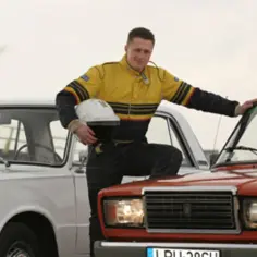 Mężczyzna w kombinezonie rajdowym opiera się o czerwonego Łada 2107, obok stoi biały Fiat 125p.