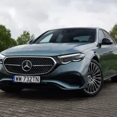 Elegancki, zielony Mercedes E220d z charakterystycznym grillem i felgami stoi na brukowanej nawierzchni, otoczony zielenią.
