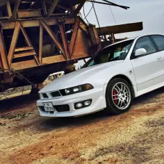 Biały Mitsubishi Galant, lśniący obok zardzewiałej, industrialnej konstrukcji.
