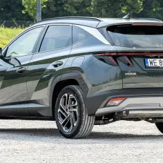 Nowy Hyundai Tucson w ciemnozielonym kolorze, zaparkowany na żwirowej drodze. Widoczny tył pojazdu z charakterystycznymi światłami i felgami.
