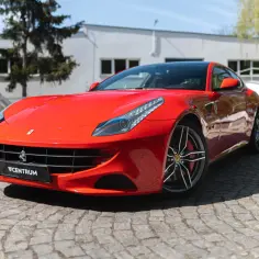 Czerwone Ferrari FF na brukowanej drodze, z budynkiem i drzewami w tle.