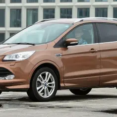 Brązowy Ford Kuga 2 z kierowcą w okularach, zaparkowany na mokrym parkingu przed nowoczesnym budynkiem.