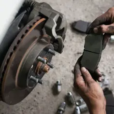 Mechanik pokazuje zużyte klocki hamulcowe przed ich wymianą. Widać tarczę hamulcową i zacisk.