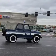 Policyjny UAZ na ulicy, z napisem "POLICJA" na drzwiach. W tle budynek z napisem "TARCZYŃSKI ARENA".
