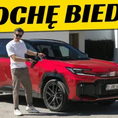 Mężczyzna w białej koszulce i brązowych spodniach stoi obok czerwonego SUV-a, wskazując na drzwi. Nad nimi żółty baner z napisem "TROCHĘ BIEDA...".