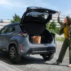 Szara Toyota Yaris Cross z otwartym bagażnikiem, w którym widać torbę. Kobieta zamyka klapę. Wymiary auta pozwalają na wygodne pakowanie.