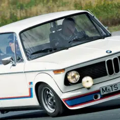 Białe BMW 2002 Turbo z kolorowymi pasami na zderzaku i błotnikach, pędzi po torze.