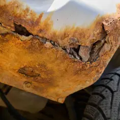 Widoczne zaawansowane ogniska rdzy na podwoziu samochodu, które niemal całkowicie zniszczyły fragment karoserii. Obok widać fragment opony.