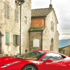 Czerwone Ferrari Italia na tle kamiennych domów i wzgórz.