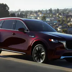 Elegancka, bordowa Mazda CX-90 pędzi po autostradzie, z miastem w tle.