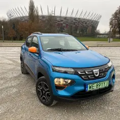 Niebieski dacia elektryk z pomarańczowymi akcentami zaparkowany na żwirowym placu przed Stadionem Narodowym w Warszawie.