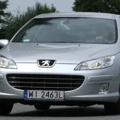 Srebrny Peugeot 407 2.0 HDI jedzie po drodze, otoczony zielenią.