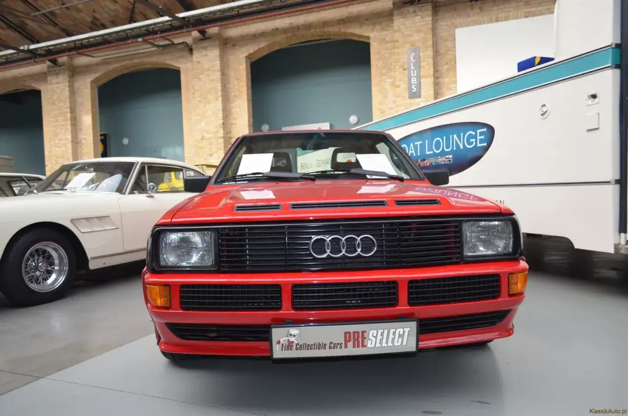 Czerwony Audi Sport Quattro z charakterystycznym grillem i logo. Obok stoi biały klasyk.