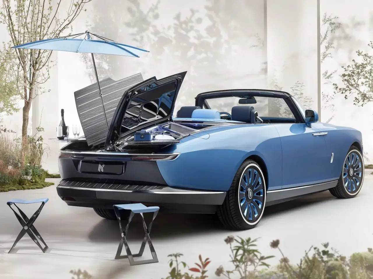 Niebieski Rolls-Royce Boat Tail z otwartym bagażnikiem, w którym znajduje się zestaw piknikowy, parasol i dwa składane krzesełka.