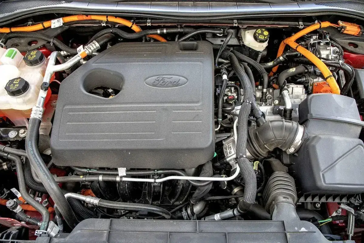 Silnik Ford Kuga 2. Hybrydowy układ napędowy z widocznymi pomarańczowymi przewodami wysokiego napięcia i czarną osłoną silnika z logo Ford.