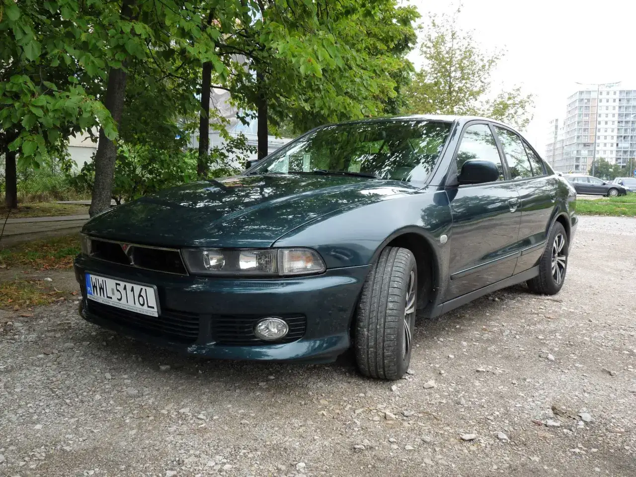 Elegancki, ciemnozielony Mitsubishi Galant stoi na żwirowej drodze, otoczony drzewami.