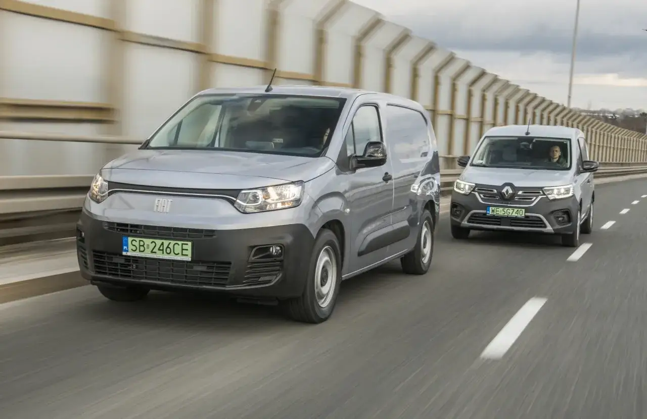 Dwa dostawcze samochody jadące po drodze. Z przodu srebrny fiat elektryczny, za nim Renault.