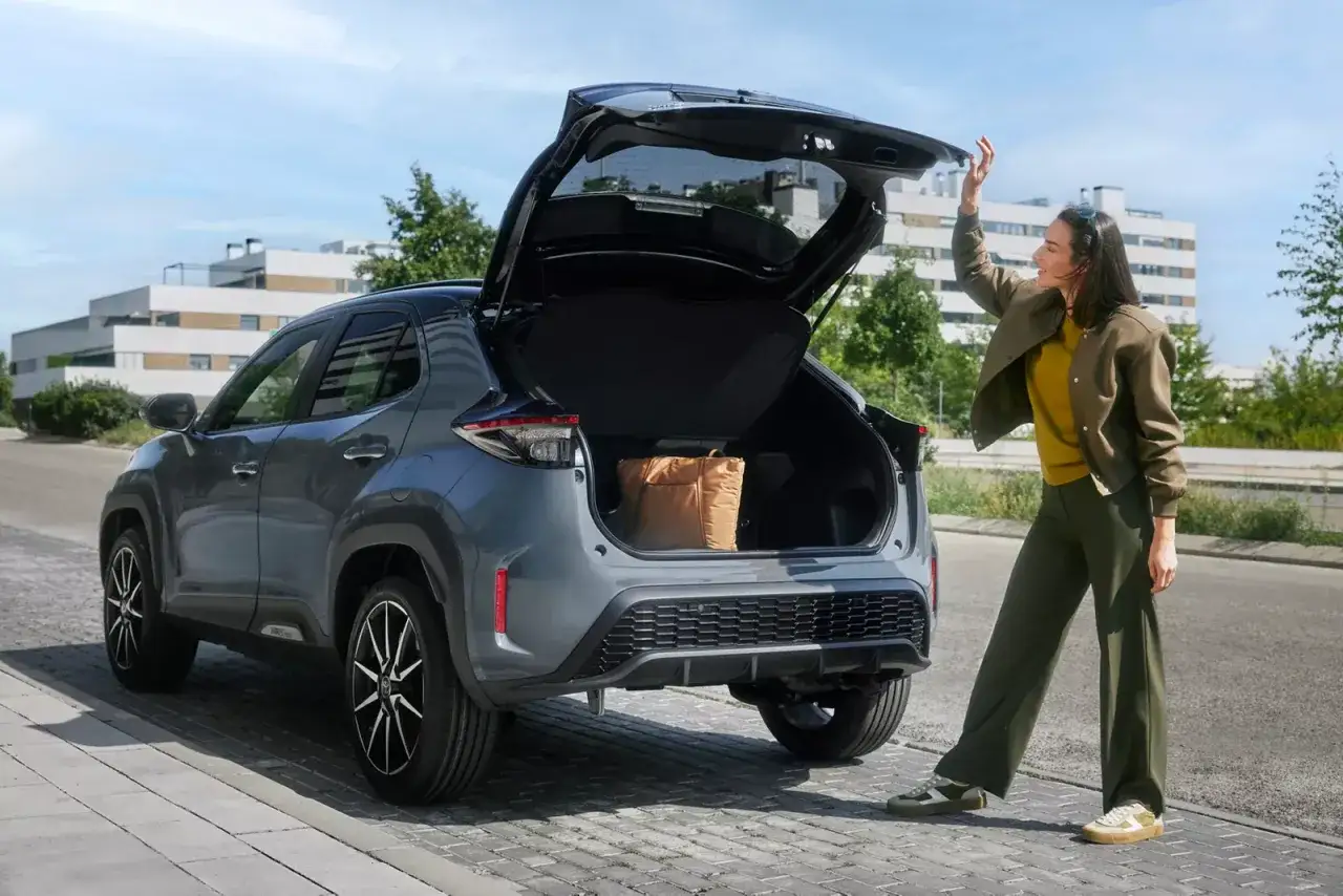 Szara Toyota Yaris Cross z otwartym bagażnikiem, w którym widać torbę. Kobieta zamyka klapę. Idealne wymiary do miasta.