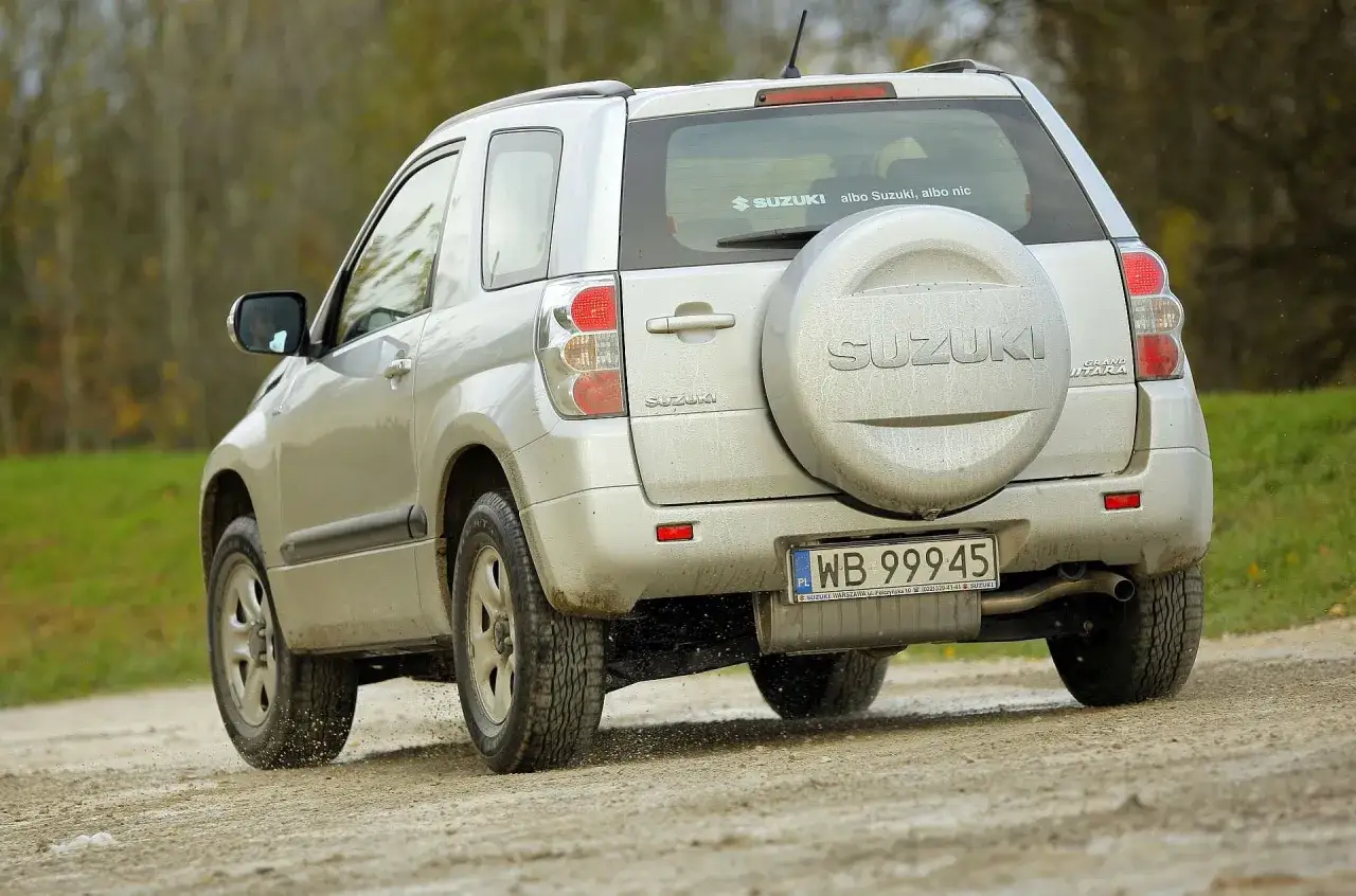 Srebrny Suzuki Grand Vitara 2 na drodze gruntowej. Zapasowe koło na tyle, naklejka na szybie.