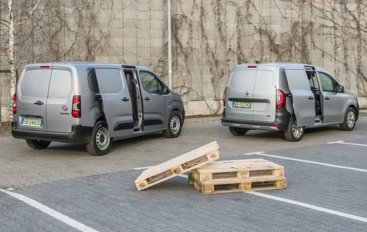 Dwa srebrne dostawcze samochody, jeden fiat elektryczny, stoją obok siebie na parkingu. Przed nimi leżą drewniane palety.